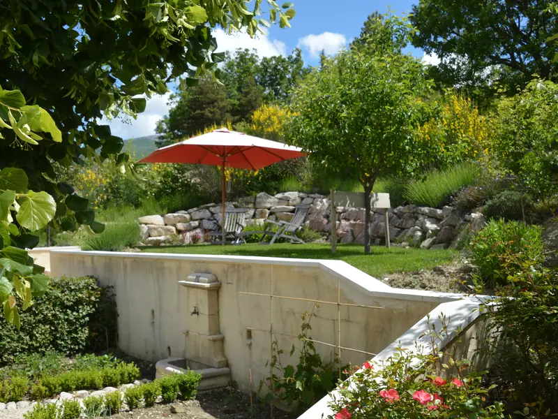 De tuin met een stenen fontein, ligstoelen onder een rode parasol en bloeiende brem